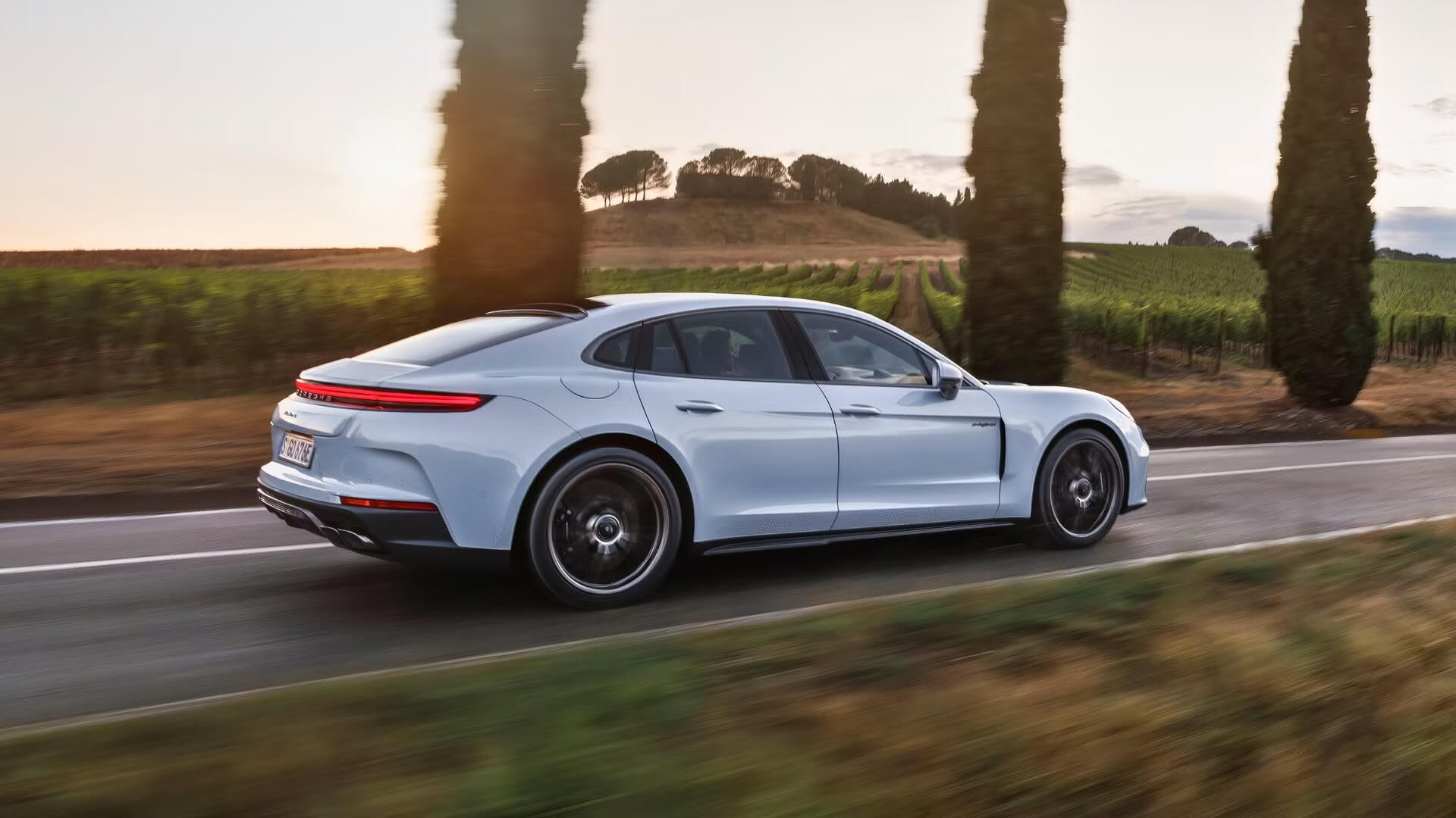 Светло-голубой Porsche Panamera Turbo S E-Hybrid 2024 года едет по дороге, окруженной сельским пейзажем с кипарисами.