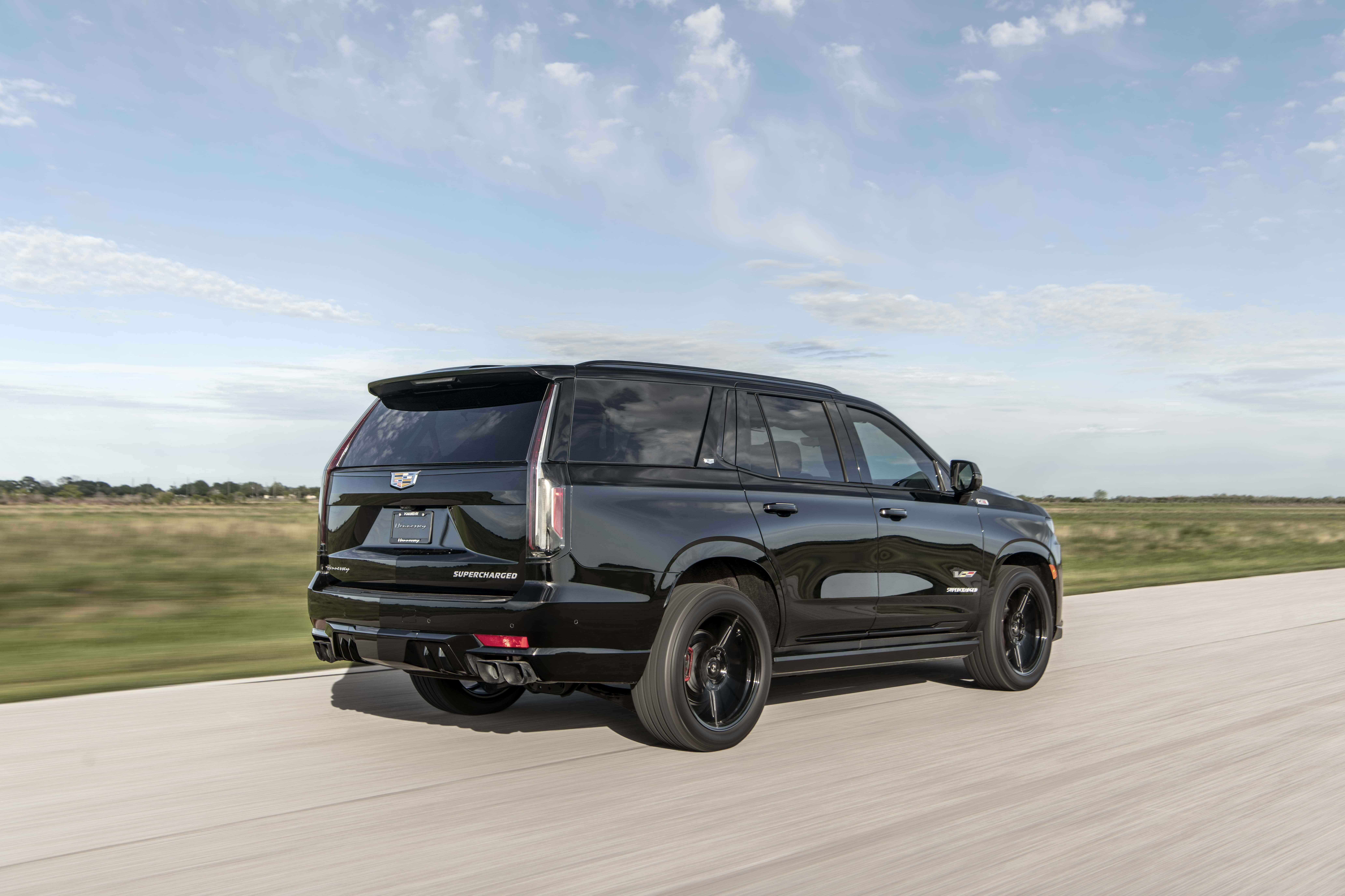 Черный Cadillac Escalade Supercharged в движении по скоростной трассе на фоне открытого неба и зелёного ландшафта.