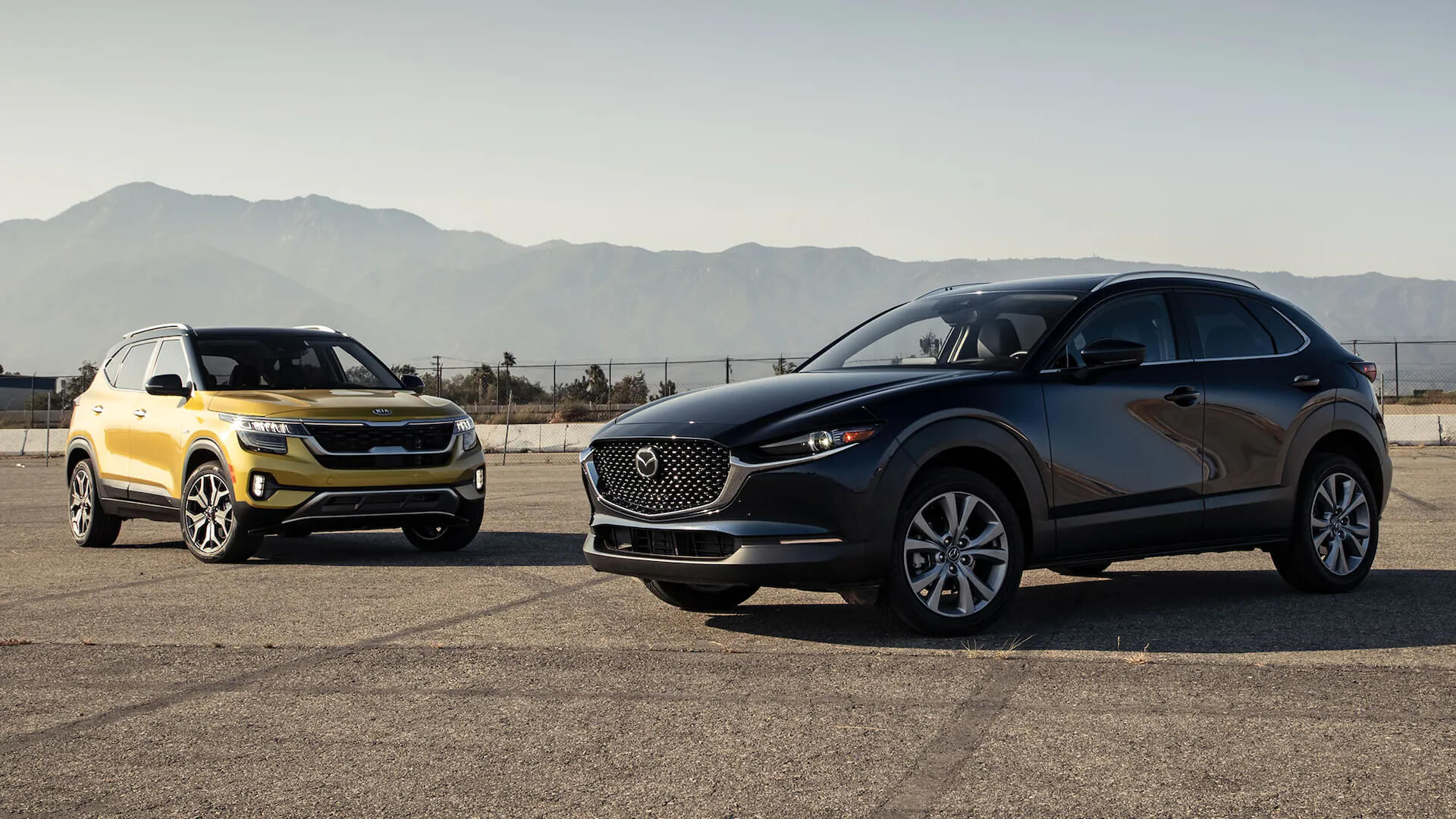 Черный Mazda CX-30 и желтый Kia Seltos припаркованы на открытой местности с горами на фоне.