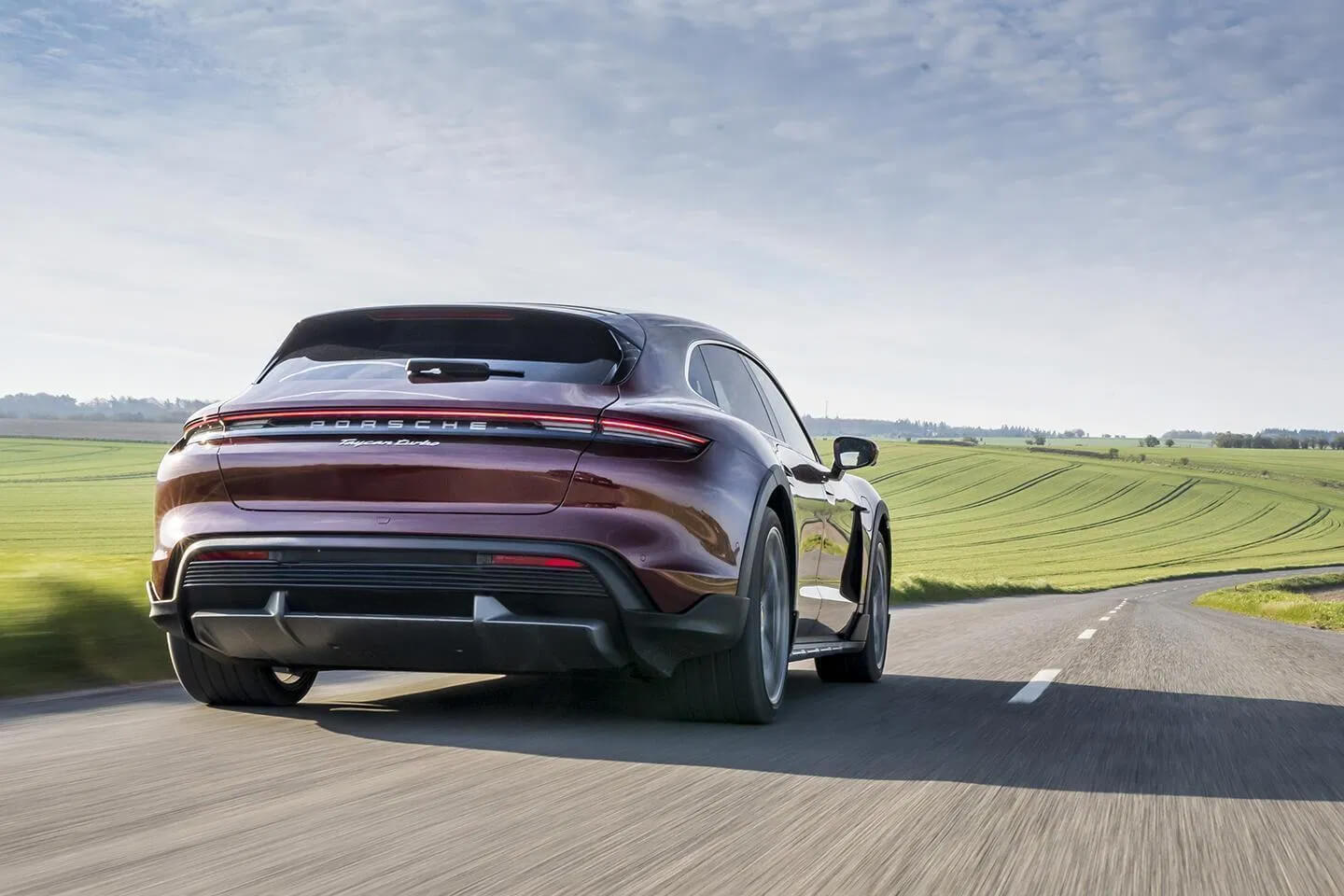Porsche Taycan Turbo S Cross Turismo: бордовый электрокар в движении на трассе с зеленым полем на фоне.