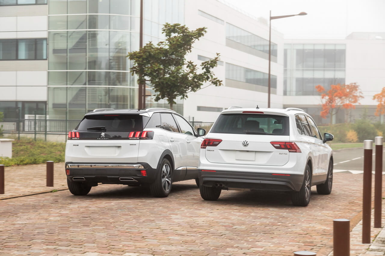 Белые Volkswagen Tiguan и Peugeot 3008 припаркованы на фоне современного здания в деловом районе.