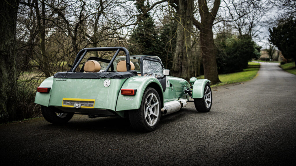 Зелёный Caterham Seven с открытым кузовом стоит на асфальтированной дороге среди деревьев. На задней части автомобиля виден номерной знак "HRZ 737". Автомобиль имеет классический дизайн с длинным капотом и короткой кабиной.